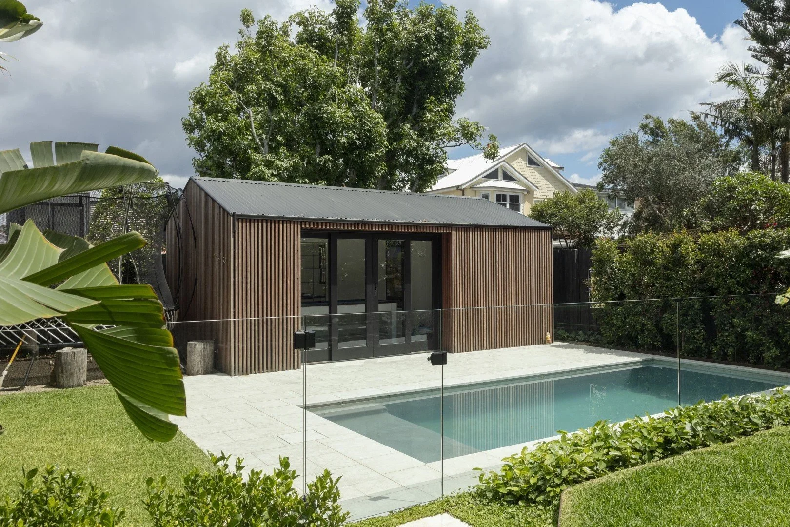 A backyard with a rectangular swimming pool, surrounded by a glass fence, and a small wooden pool house with a metal roof in the background. There are lush trees and bushes around the area, with a partly cloudy sky overhead.