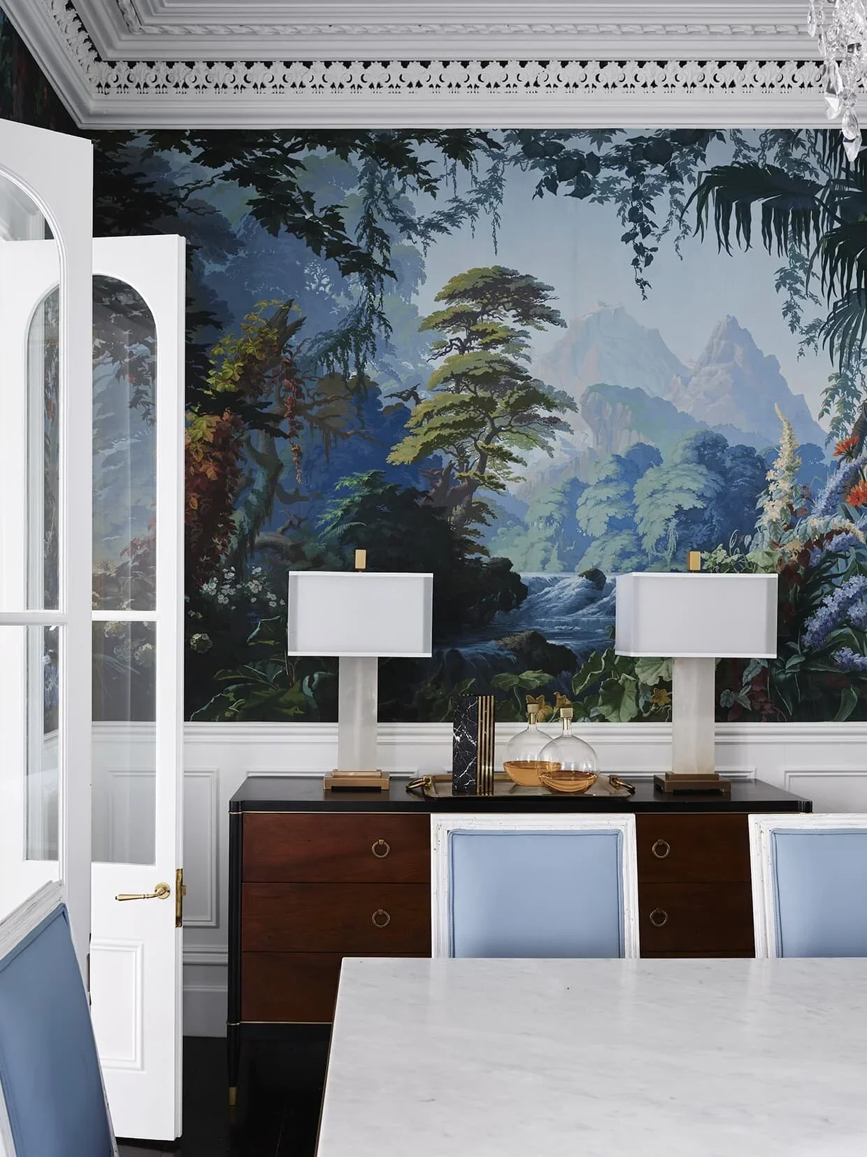 Decorative interior room with a large nature-themed mural featuring trees, mountains, and a river, white and blue chairs, a black and brown sideboard with two table lamps, and a marble-topped table.