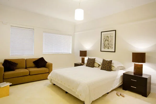 A bedroom with a beige couch, four pillows, a bed with white bedding, two dark nightstands with table lamps, a piece of framed art above the bed, and two windows with blinds.