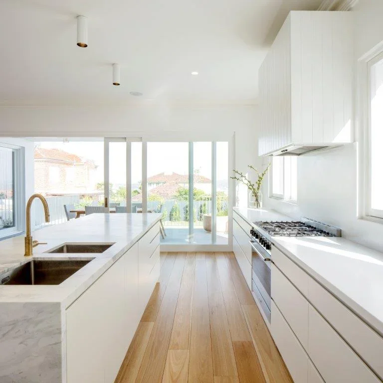 Bright, modern kitchen with white cabinets, a marble island with a gold faucet, and large windows offering a view of a patio and neighborhood.