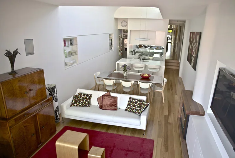 Living room with white sofa, wooden cabinet, wall decor, dining table with chairs, open kitchen, wood flooring, wall-mounted TV, and decorative items.