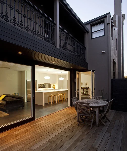 Photo of a modern two-story house with a balcony, large glass sliding doors, and a wooden outdoor deck with an outdoor dining table and chairs.