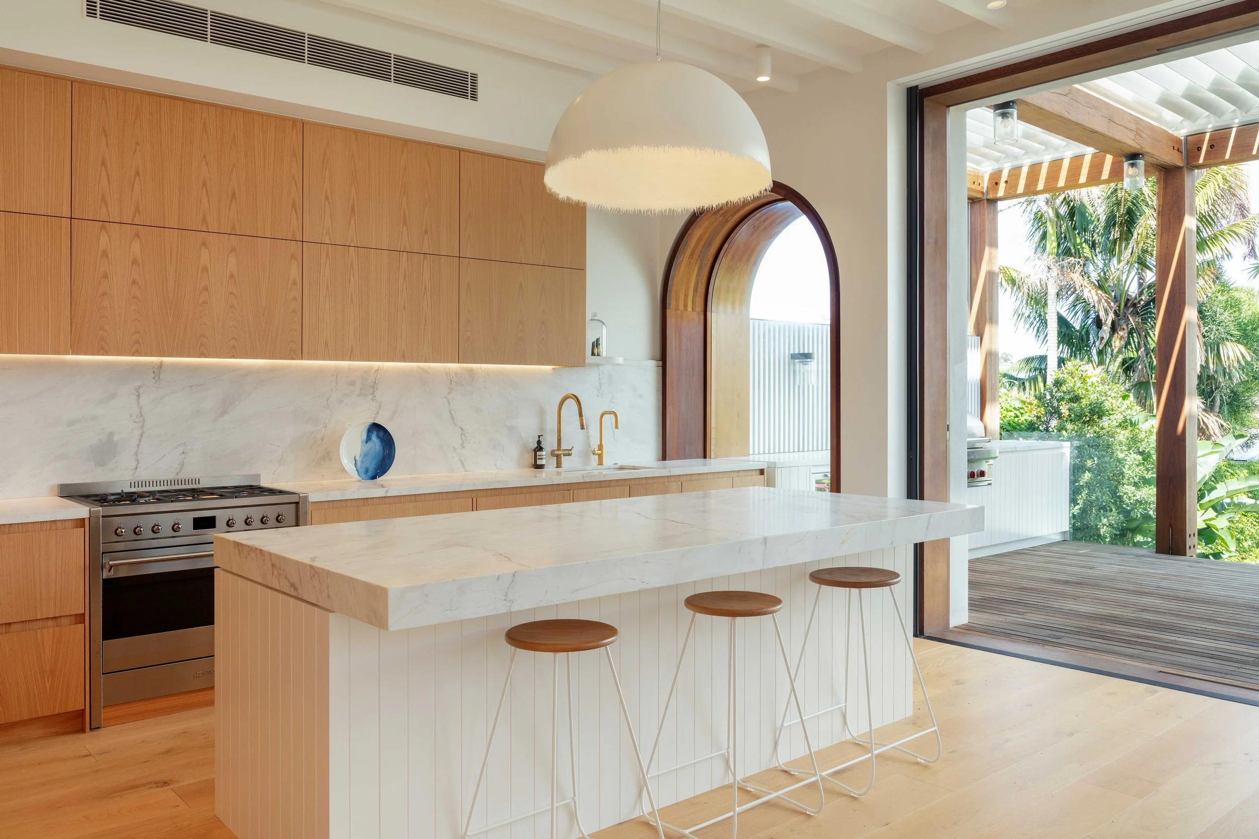 A contemporary kitchen within the Bronte House project by To The Mil, defined by natural timber cabinetry, a large marble island and an elegant arched opening connecting the interior to the outdoor terrace.