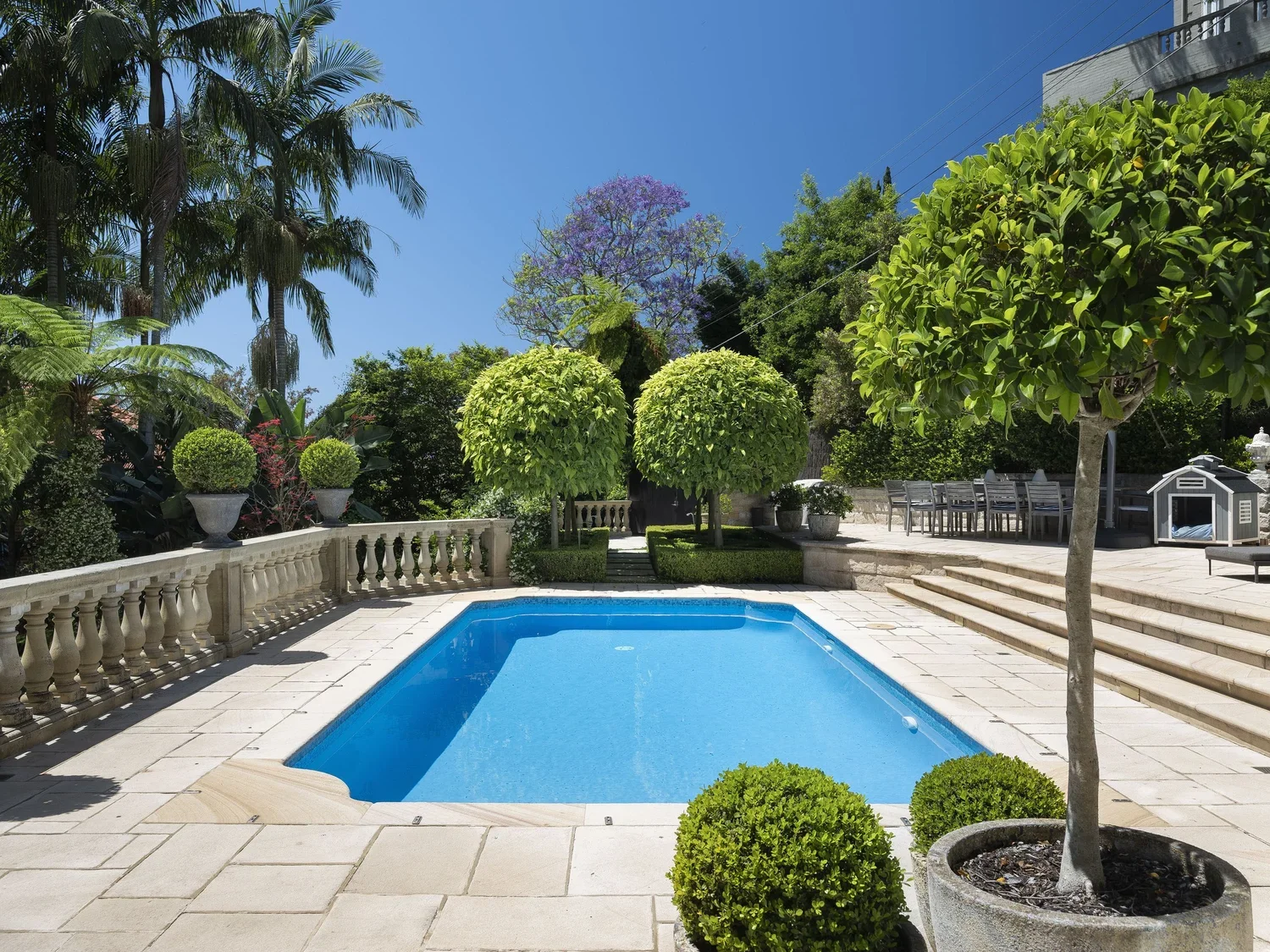 Backyard with blue swimming pool, surrounded by stone patio, with green trees and plants, including potted plants, hedges, and tall palm trees, under a clear blue sky.