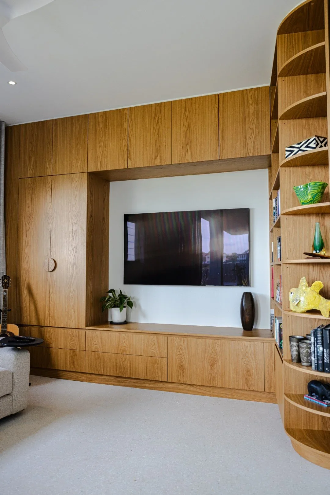 Living room with built-in wooden entertainment center, wall-mounted flat-screen TV, a potted plant, decorative vases, and a curved wooden bookshelf with books and decorative items.