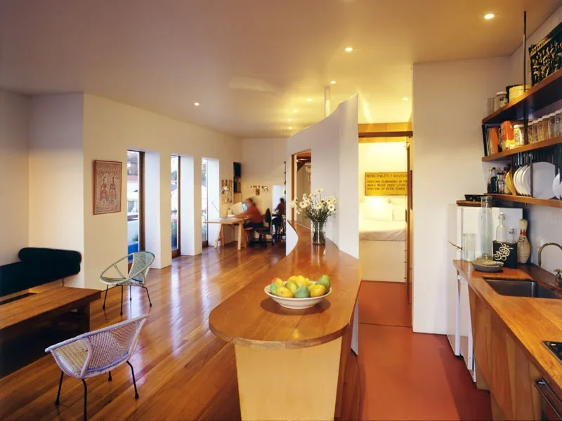 Interior of a cozy cafe with wooden floors, a curved wooden counter with a bowl of fruit, shelves with plates and jars, and customers sitting at a table.