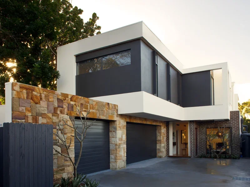Modern two-story house with white and black exterior, stone accents, and large garage doors, surrounded by trees.