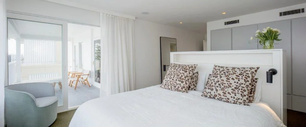 Modern hotel room with a white bed, leopard-print pillows, a vase of white flowers, and a balcony with outdoor seating.