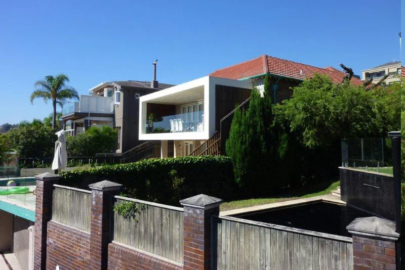 Modern house with a white exterior and large balcony, surrounded by green trees and a brick fence.