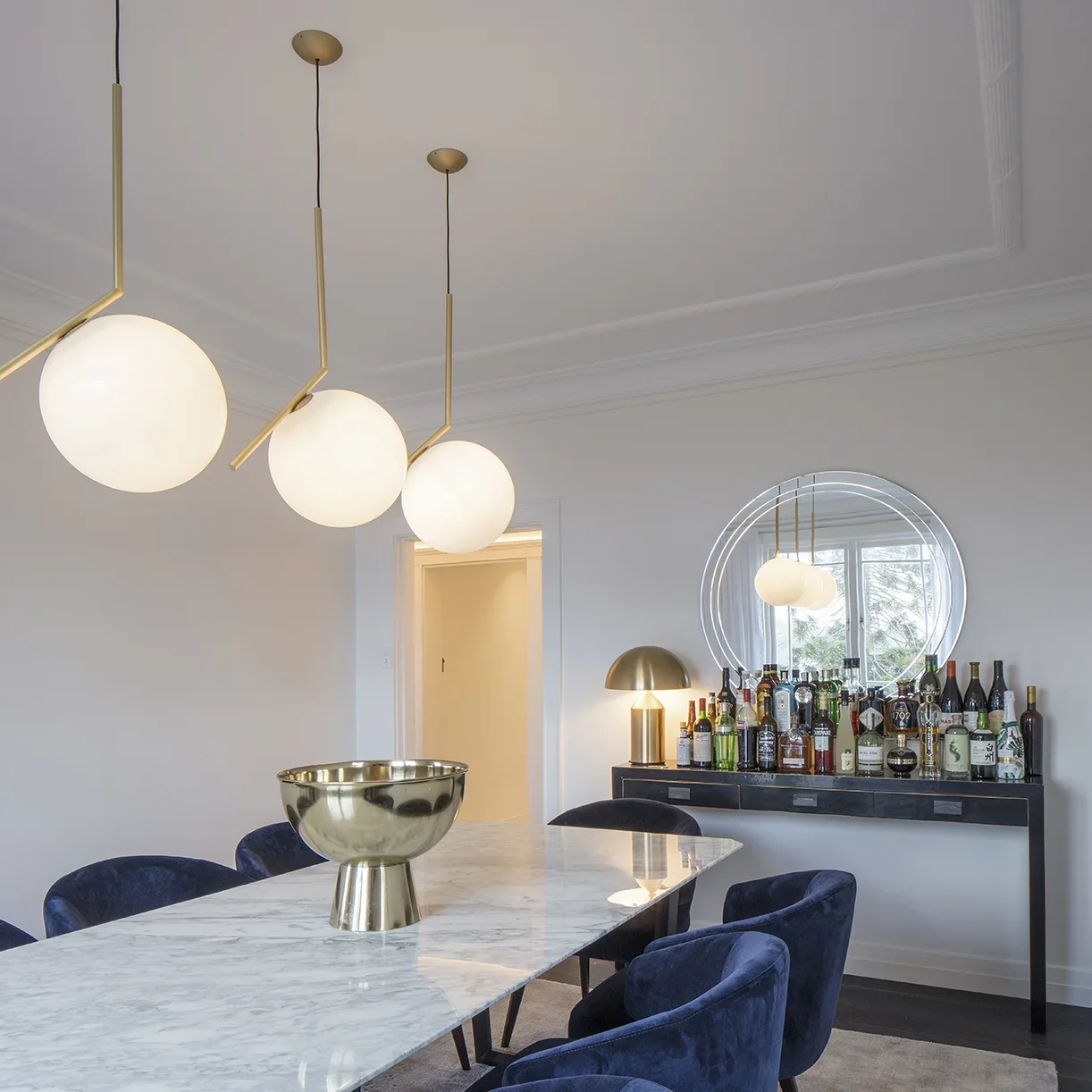 Modern dining room with a marble table, navy blue chairs, contemporary pendant lights, a bar cart with liquor bottles, and a round mirror reflecting the window outside.