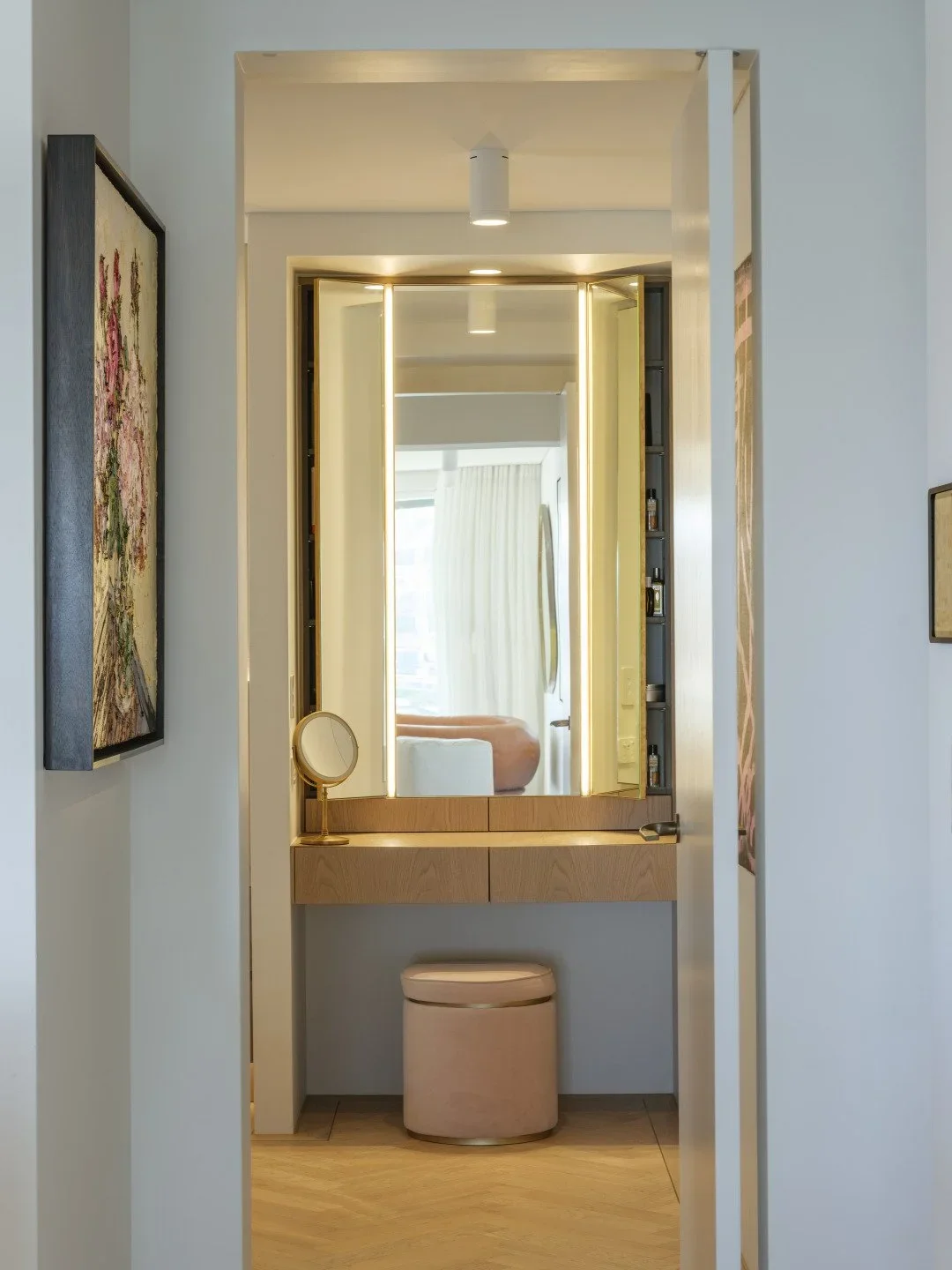 A modern vanity area with a large illuminated mirror, a small round mirror, and a pink cushioned stool, reflecting a nearby bed and curtains.