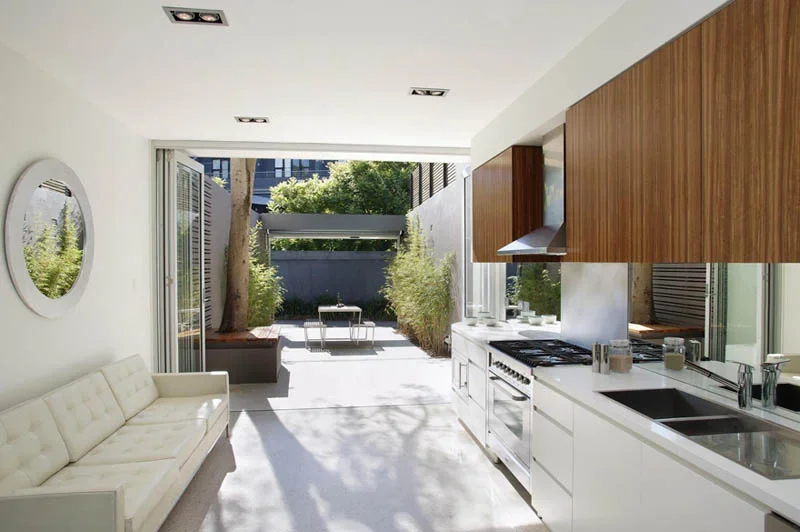 Modern open-concept kitchen and patio with white and wooden cabinetry, stainless steel appliances, a white couch, and outdoor seating facing a landscaped garden.