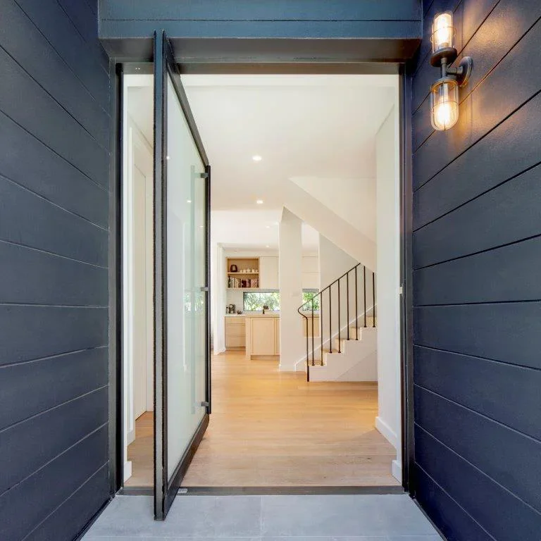 Open glass door leading into a modern house interior with wooden flooring, white walls, a staircase with black railing, and a view of the kitchen and living area.
