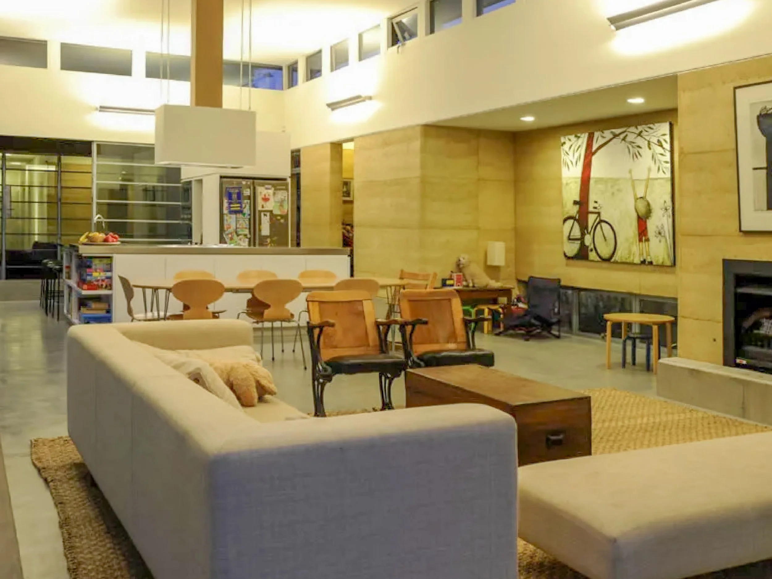 A spacious, cozy living area with beige sofas, wooden chairs, a coffee table, and a fireplace. There is a large wall art of a bicycle and trees, and a dining area with a table and chairs. The room has high ceilings and ample natural light.