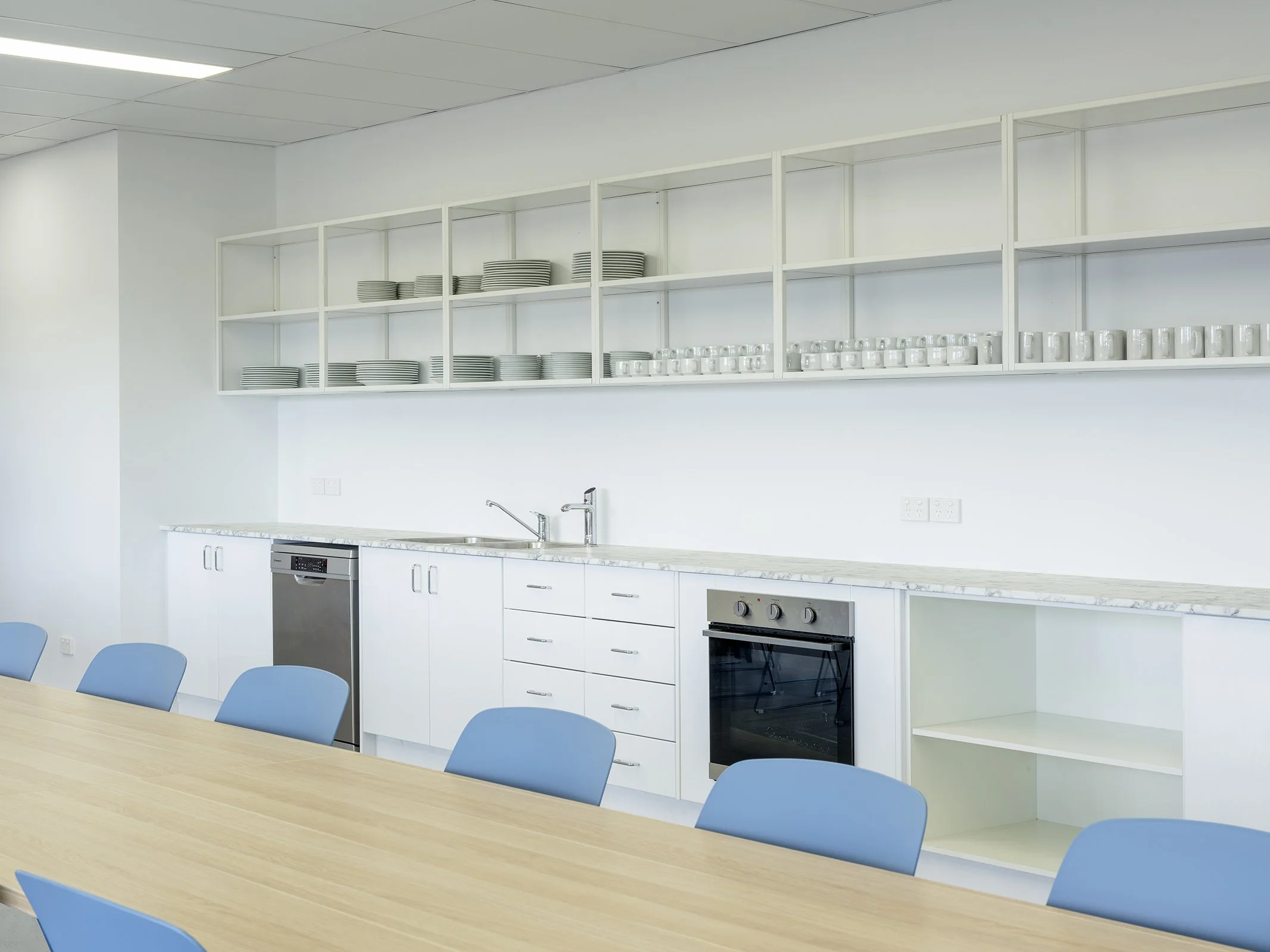 Modern kitchen with white cabinets, open shelving with white dishes, blue chairs around a wooden table, stainless steel appliances including oven and dishwasher, white walls, and a marble countertop.