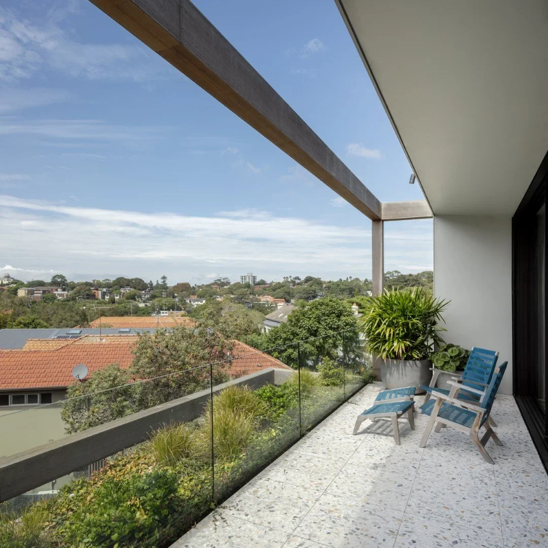 View from a balcony showing a cloudy sky, trees, and houses with red-tiled roofs, two blue and white outdoor chairs, and a large potted plant.