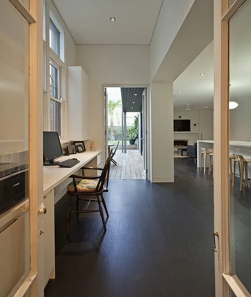 Interior view of a home with a small workstation on the left, including a computer and framed photos, leading to a deck with outdoor seating and greenery in the background.