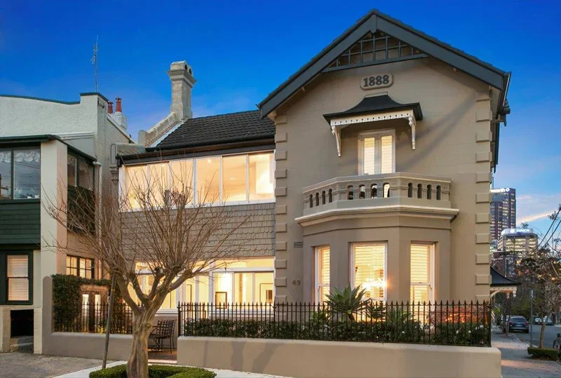 A historic two-story house built in 1888 with a beige exterior, gabled roof, and front porch with balustrade, next to a modern building with large windows.