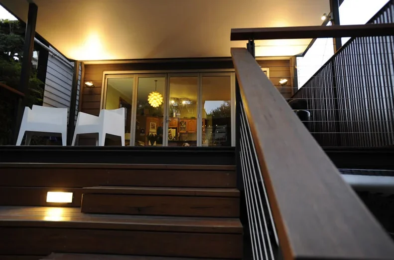 Exterior view of a house with sliding glass doors, wooden stairs, and patio balcony, with outdoor chairs and interior visible through the doors.