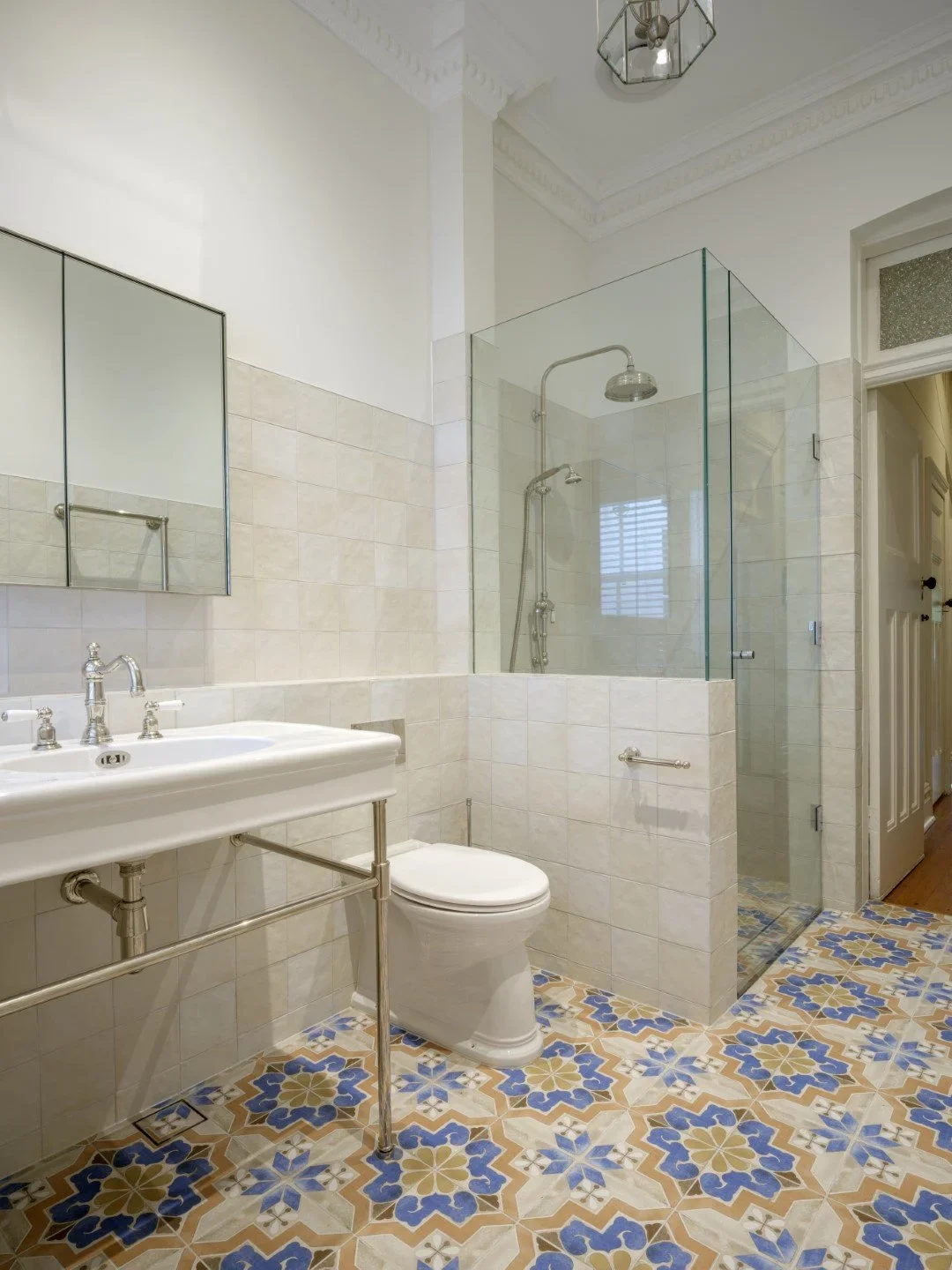 A bathroom with a white sink, toilet, and a separate shower enclosed in glass. The floor has patterned tiles, and the walls have beige tiles with a mirror above the sink. The ceiling has intricate molding, and there's a ceiling light fixture.