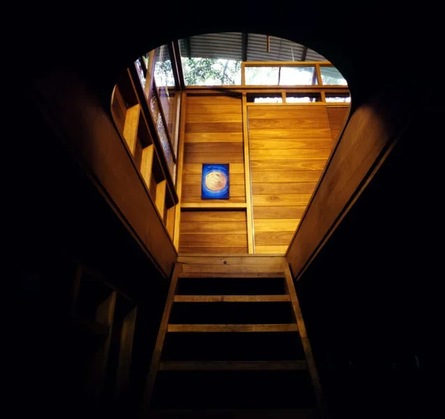 Looking up a wooden staircase toward a circular opening leading to a sunlit upstairs area with wood-paneled walls and a small framed picture of swirling colors.