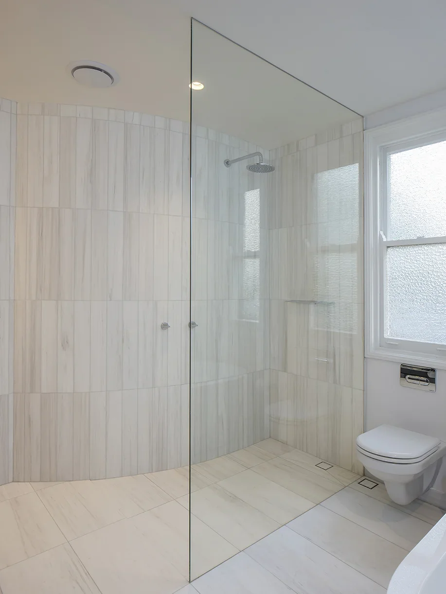 Modern bathroom with a walk-in glass shower, white marble tiles, and a wall-mounted toilet next to a frosted window.