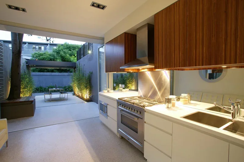 Modern kitchen with white countertops, wooden cabinets, stainless steel stove and range hood, and a sink. Looks out onto a small outdoor patio with greenery and outdoor seating.