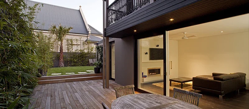 Modern outdoor patio with wooden deck, outdoor chairs, and a view into a well-lit living room with a fireplace and black sofa.