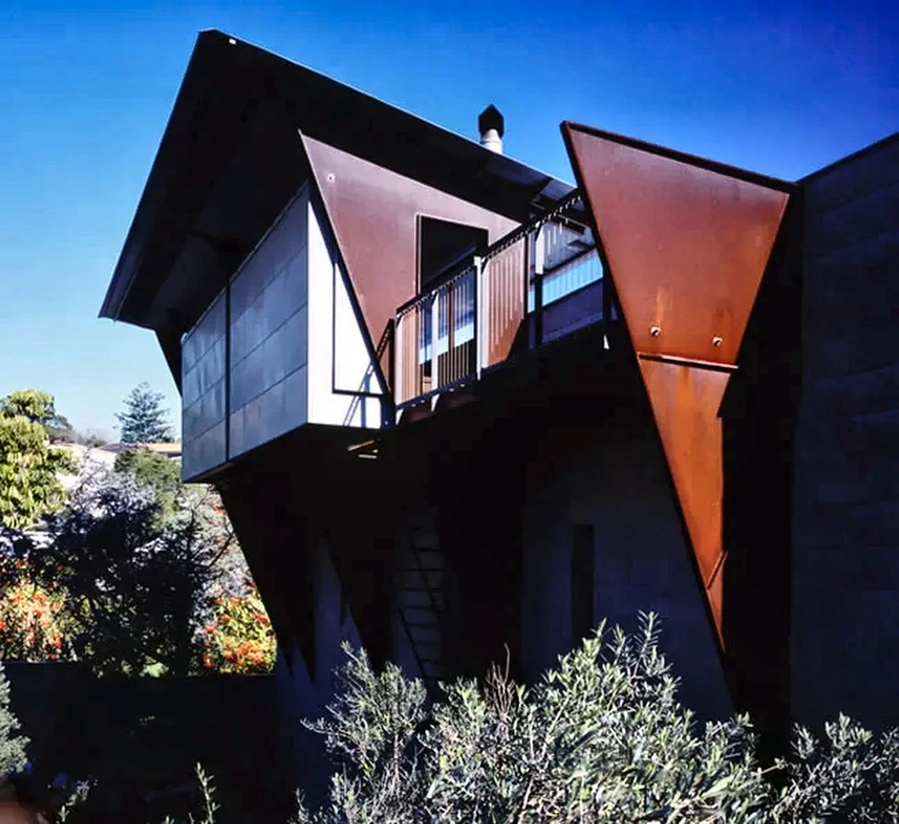 Designed by Peter Stutchbury. Modern house with angular design features rust-colored and black panels, with large windows and a balcony, set against a clear blue sky with surrounding greenery.