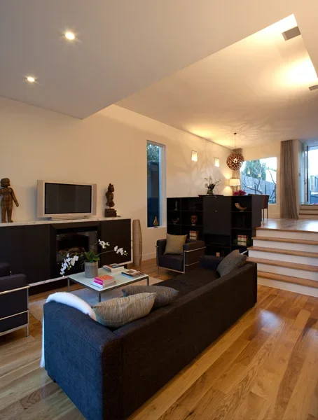 Living room with black sofa, coffee table, TV, armchair, built-in shelves, and staircase with wooden stairs, illuminated by ceiling lights and natural light from windows.