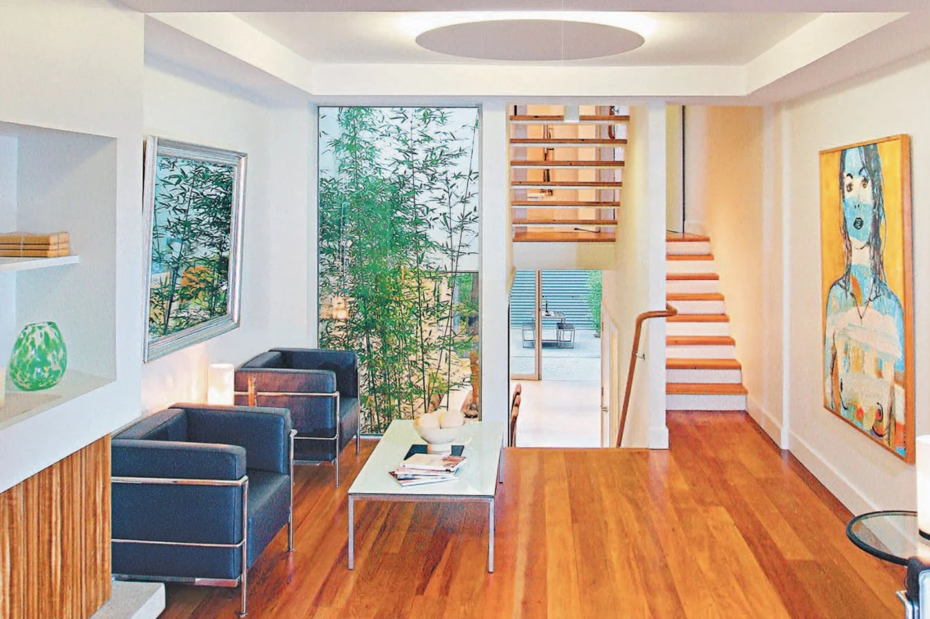 Modern living room with large window showing green bamboo, blue seating area, art on the walls, and wooden staircase.