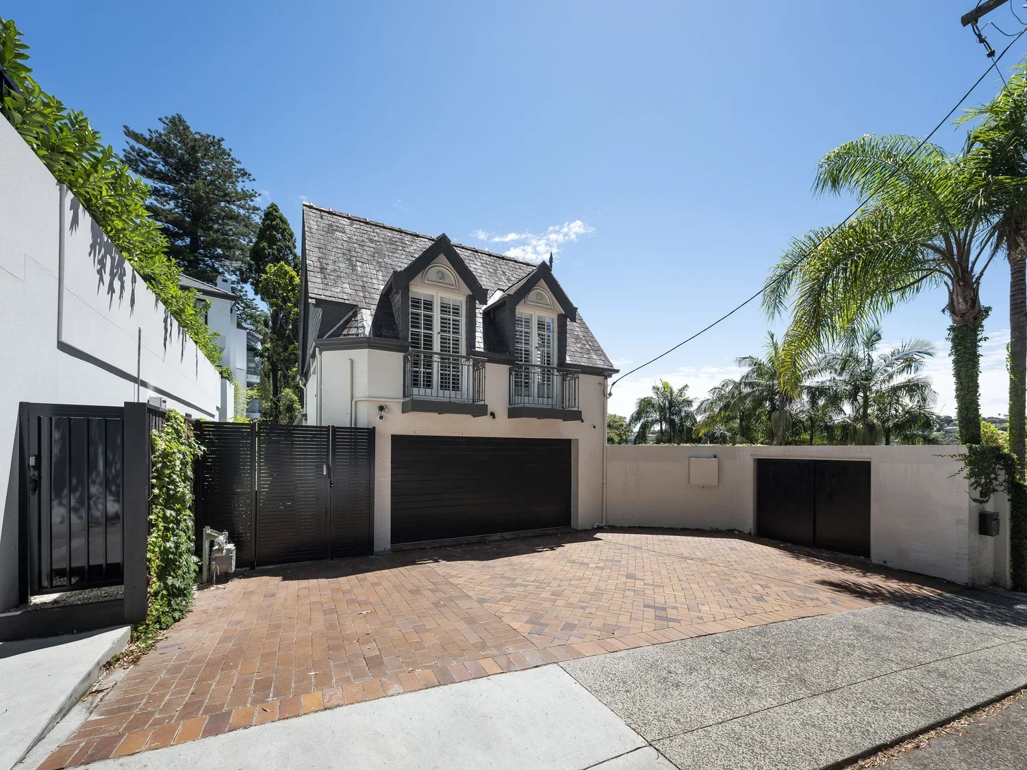 A French-style house with dormer windows and a black garage door, surrounded by a white wall, palm trees, and a brick driveway.