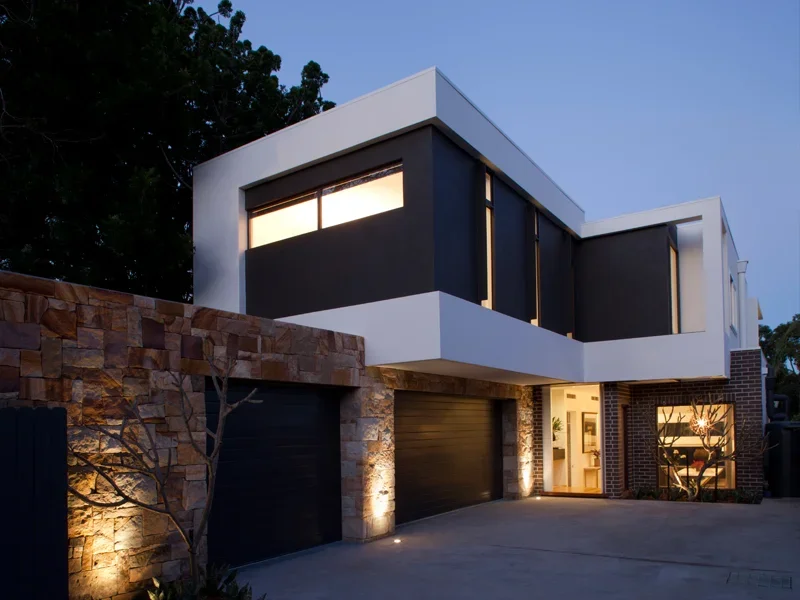 Modern two-story house with black and white exterior, large windows, stone accents, and garage doors, illuminated at dusk.