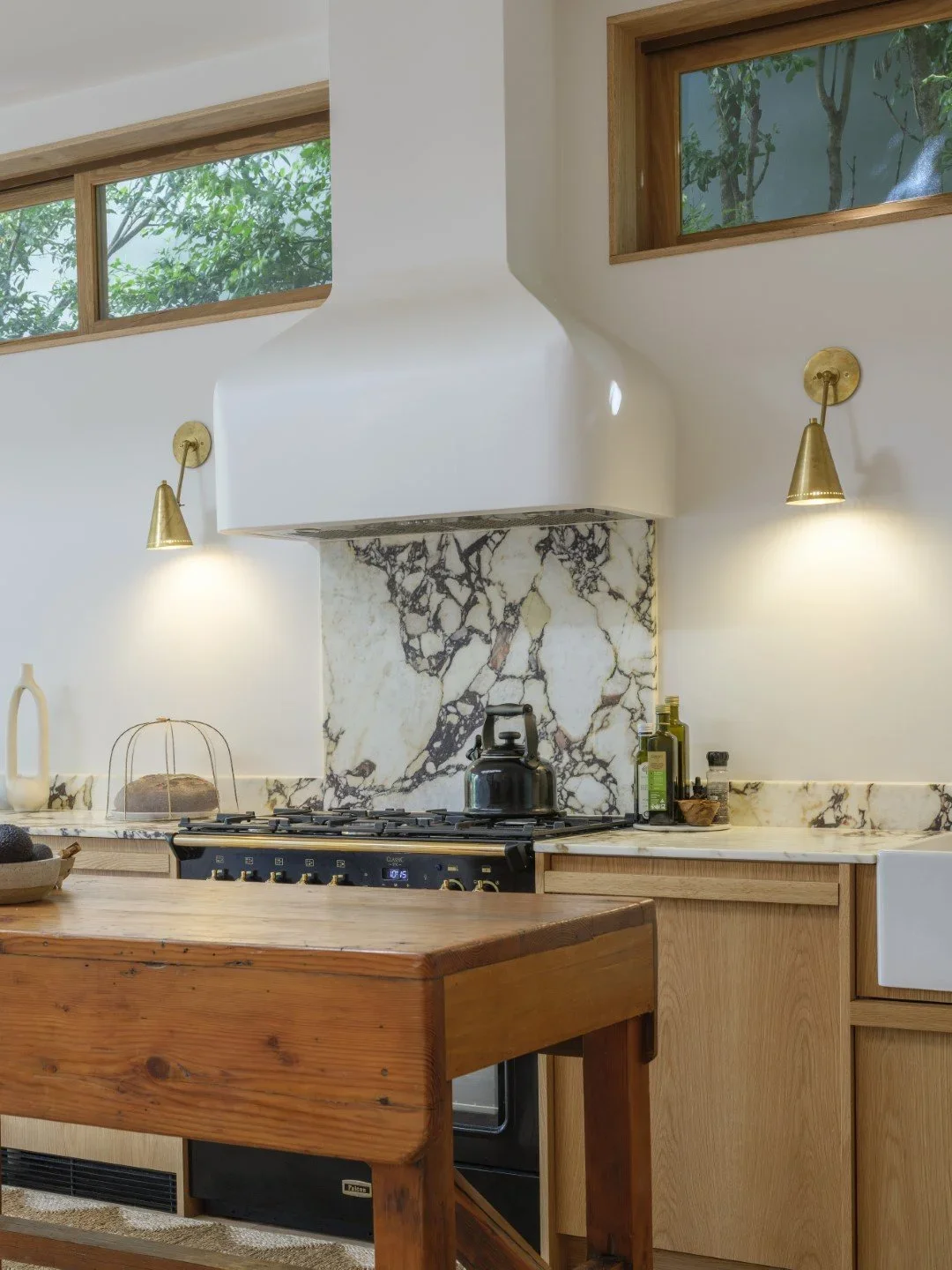 Modern kitchen with marble backsplash, wooden cabinets, black stove, brass wall-mounted lamps, and large windows with a view of trees.