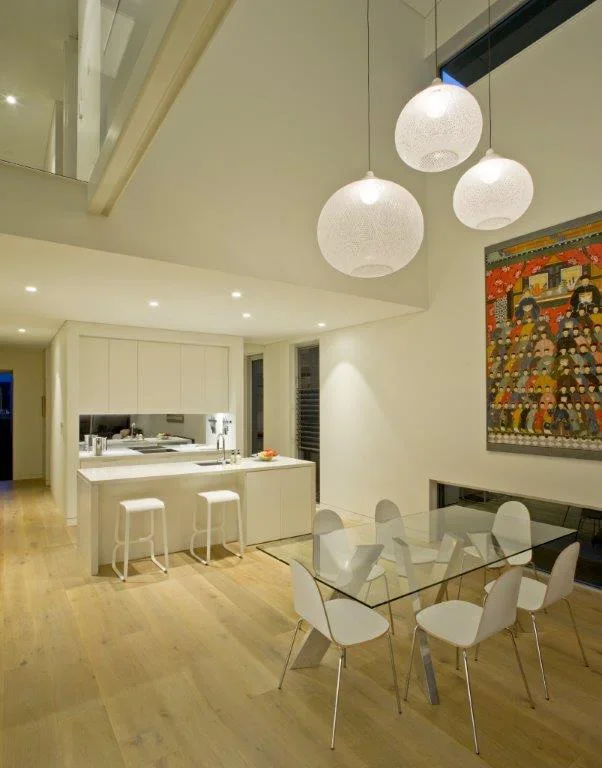 Modern dining area with a glass table and six white chairs, hanging pendant lights, and a white kitchen in the background with a stool counter.