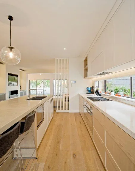 Modern kitchen with white cabinetry, a large island with seating, and stainless steel appliances. Bright room with natural light from large windows and wooden flooring.