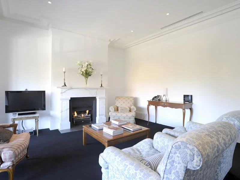 Living room with a fireplace, armchairs, a coffee table with books, a flat-screen TV, and a side table with a telephone, all decorated in a classic style with white walls and a dark carpet.