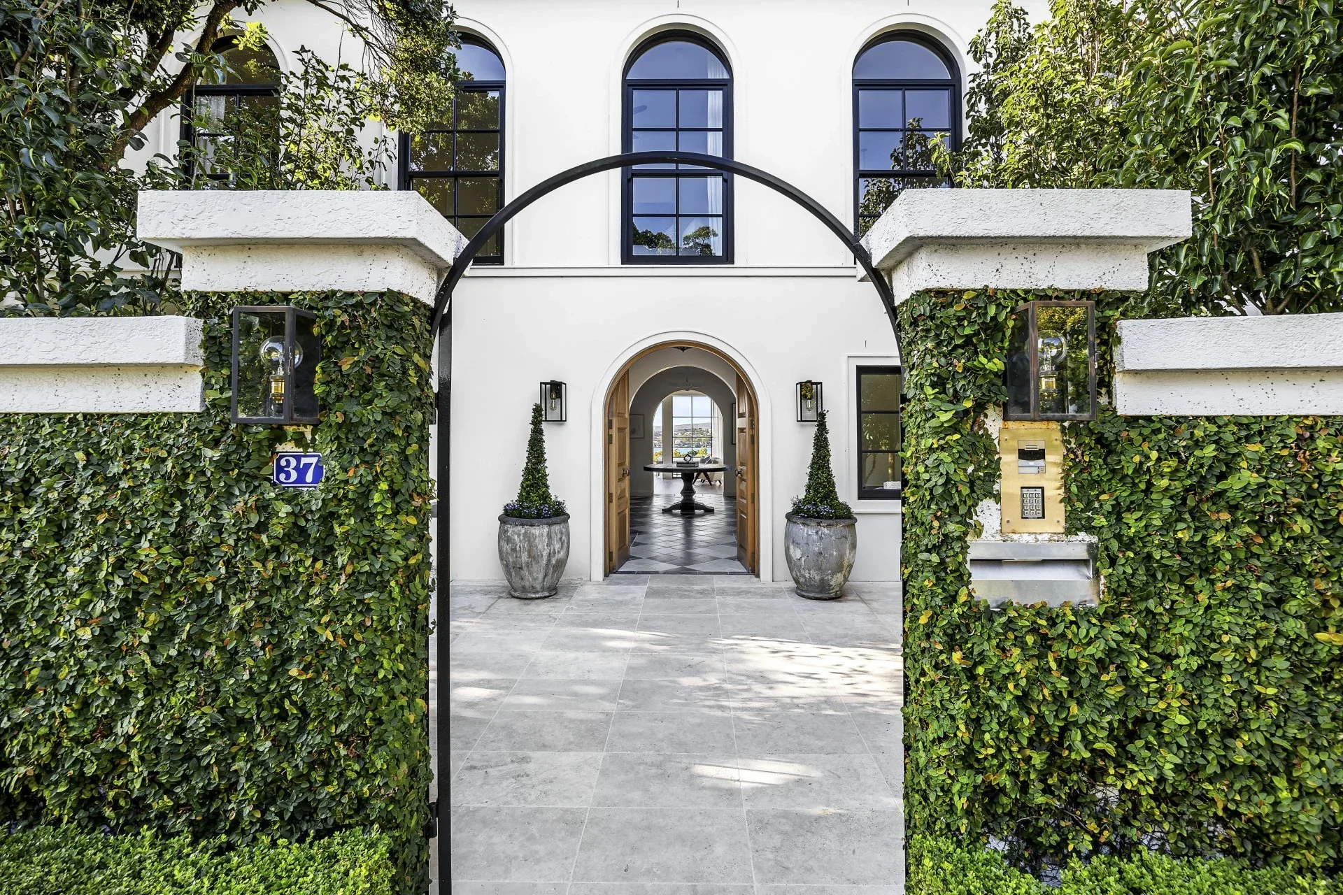Entrance to a modern white house with black window frames, a black arched gate, and plants in large concrete pots.