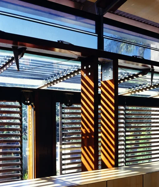 Interior view of a modern building with wooden and metal structures, large windows with horizontal blinds, and sunlight casting striped shadows on the features.