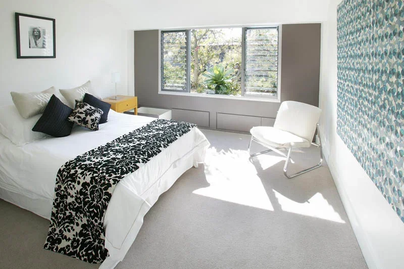 Bright bedroom featuring a bed with black and white bedding, a yellow nightstand, a white chair, a large window, and a textured blue and green wall hanging.