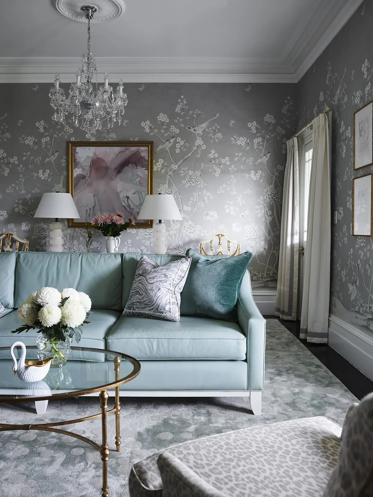 Elegant living room with a light green sofa, decorative pillows, a glass coffee table with a flower arrangement, white curtains, floral wallpaper, and a crystal chandelier.