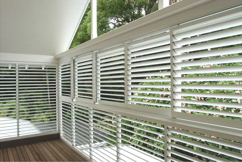 Interior of a sunroom with white wooden plantation shutters and a wooden floor, with greenery visible outside.