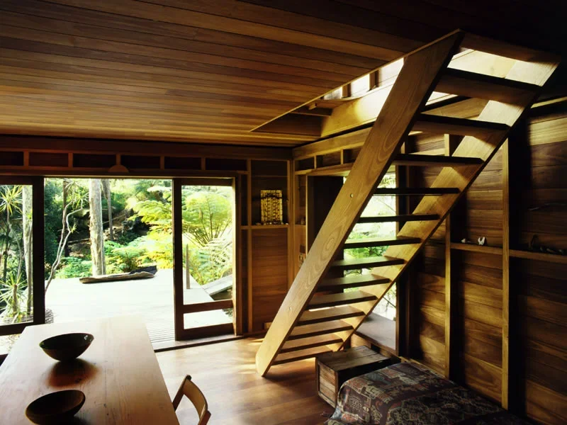 Interior view of a wooden cabin with a staircase leading to an upper level, large sliding glass doors opening to a lush green outdoor garden, and a wooden table with bowls on it.