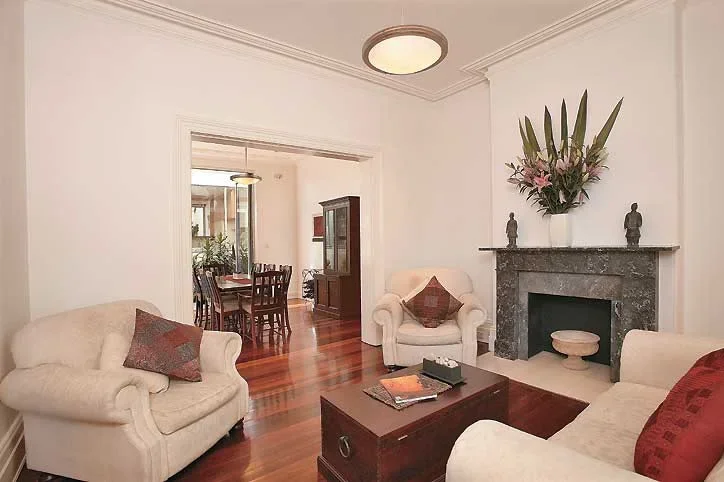 Living room with white walls, hardwood floors, white sofas, a coffee table, a marble fireplace with a flower arrangement on mantel, plants on a side table, and a dining area visible through an open doorway.