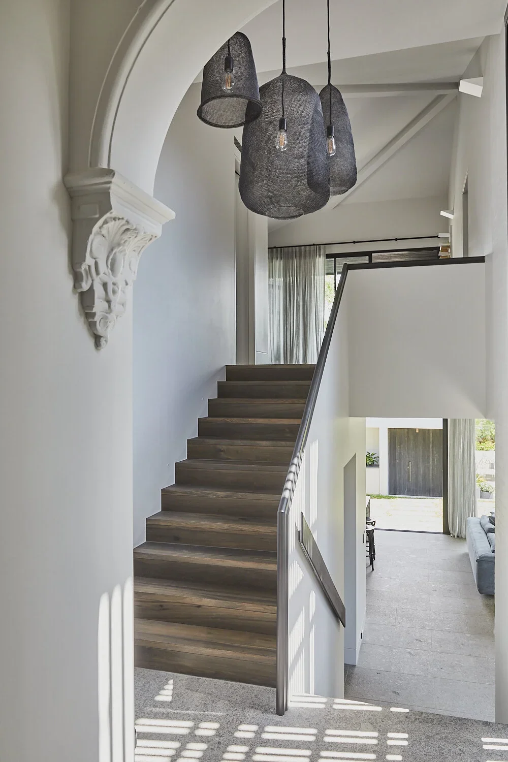 Modern staircase with wooden steps, white walls, and black pendant lights hanging from the ceiling. There is an ornate white corbel on the wall near the staircase. The area beyond the staircase has large windows with sheer curtains, allowing natural 