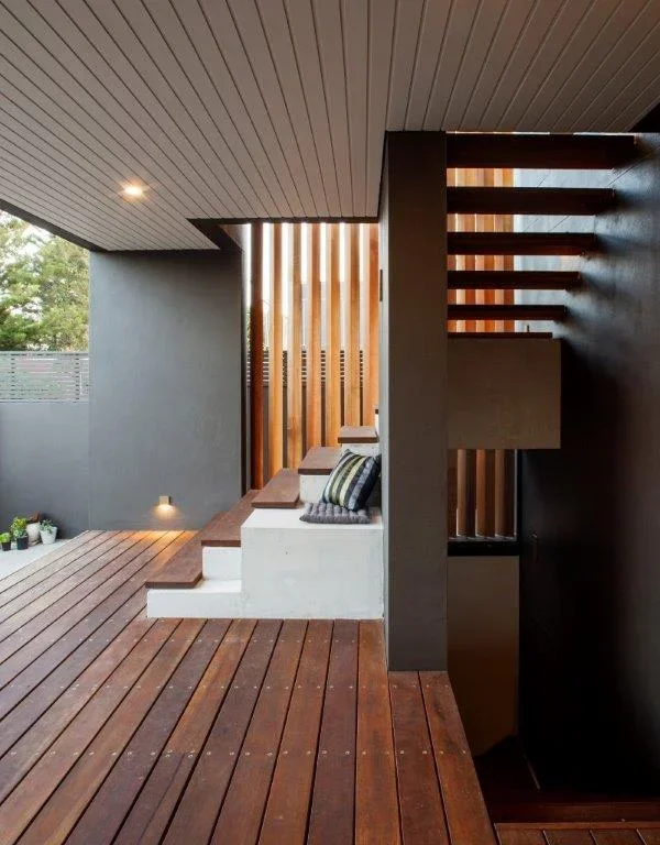 Modern outdoor patio with wooden flooring, a built-in concrete bench with cushions, and a wooden slatted privacy screen. A staircase with wooden steps and a dark wall are also visible.