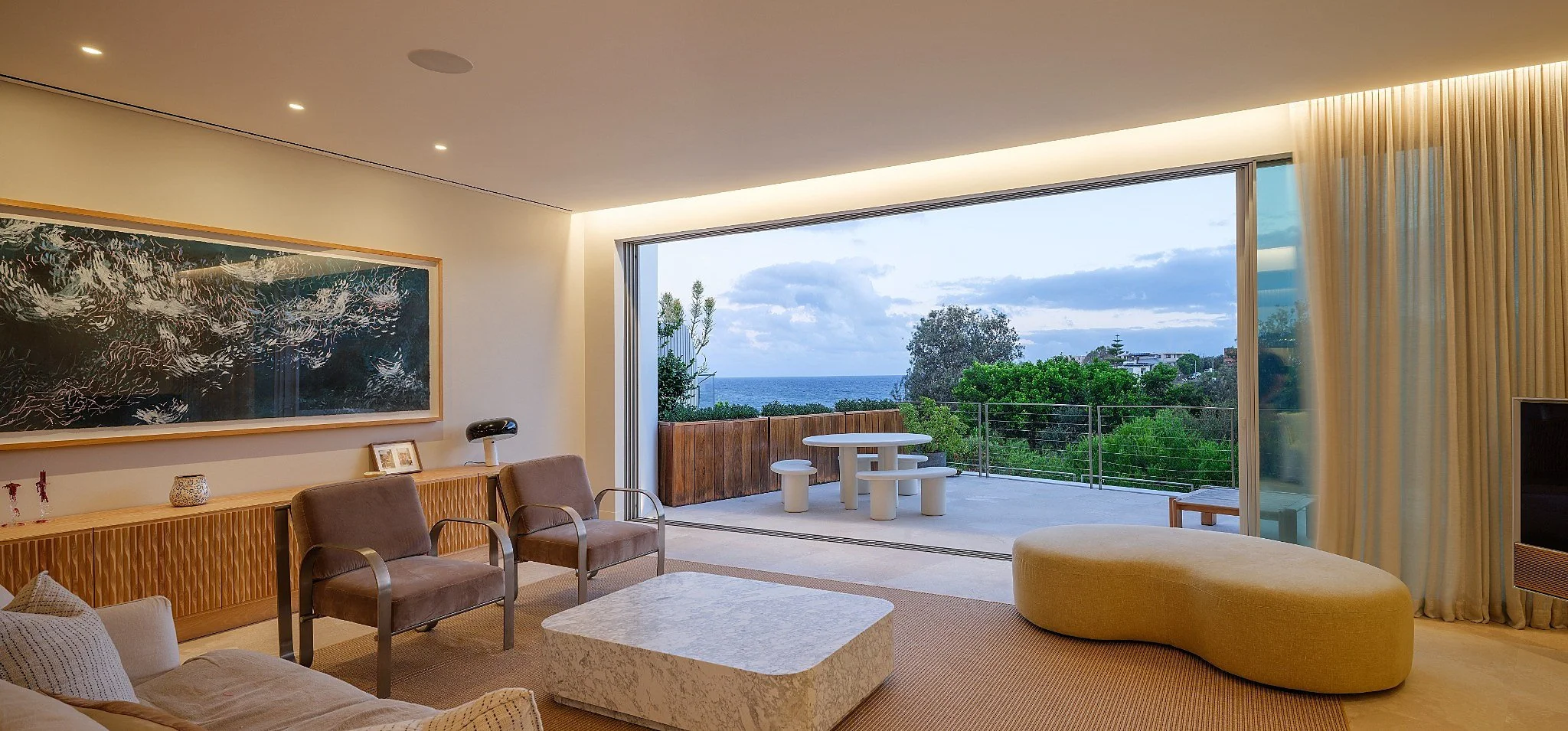 Living room with cream-colored sofa, marble coffee table, white armchair, sliding glass door showing ocean.