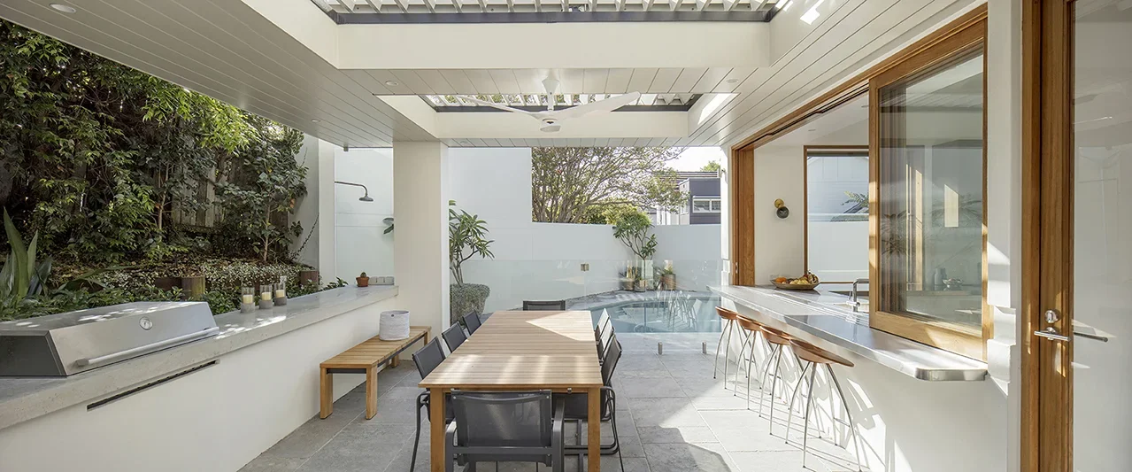 Outdoor patio with a dining table, bar area, pool, and greenery.