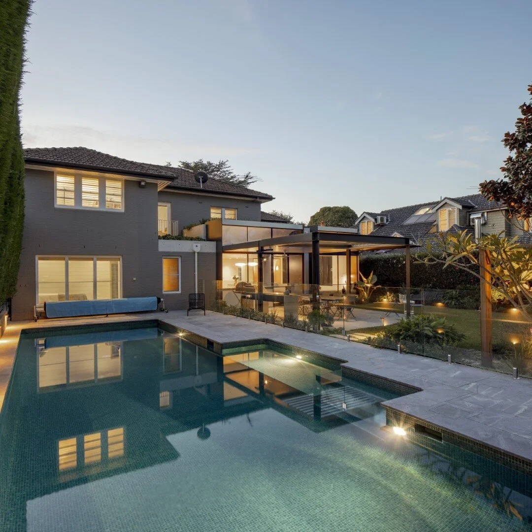 A modern backyard with a swimming pool, outdoor seating area, and a two-story house with large windows at dusk.