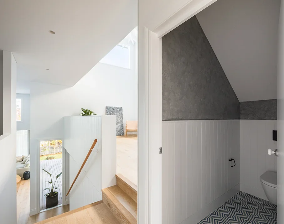 Interior view of a modern home, showing a stairway leading down to a living area with large windows and white walls, and an adjacent small room with a sloped ceiling, patterned floor tiles, and a toilet.